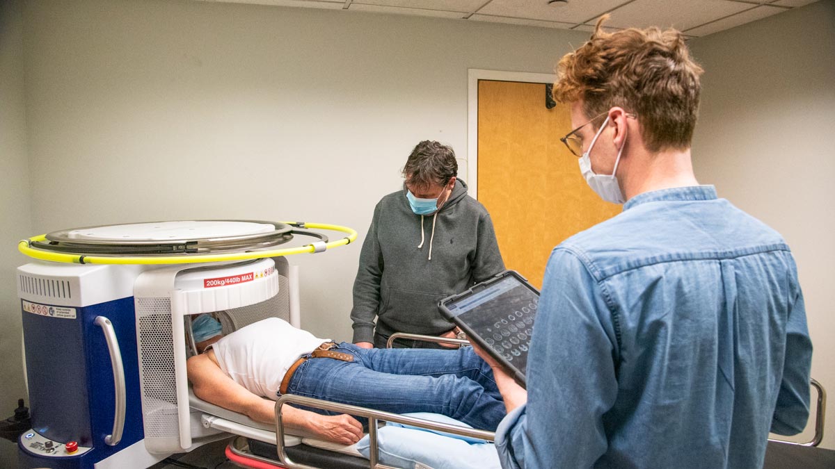 Photo: two researchers standing next to a person undergoing an ultra-low-field MRI on a Hyperfine Swoop scanner.