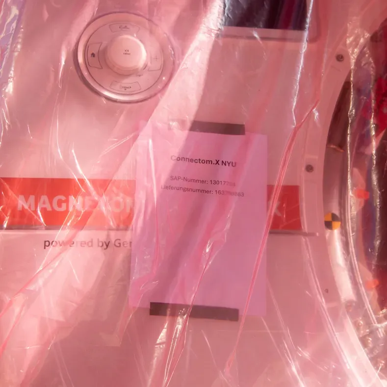 Photo: closeup showing through a translucent pink plastic cover the front face of the scanner with a taped-on sheet of paper that reads "Connectom.X NYU."